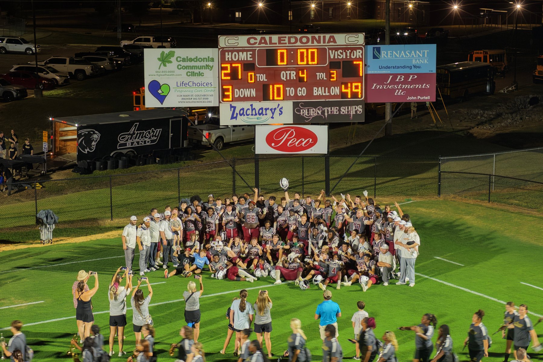 Caledonia's players and coaches pose on the field after the game, under the scoreboard showing 27 to 7 with no time left